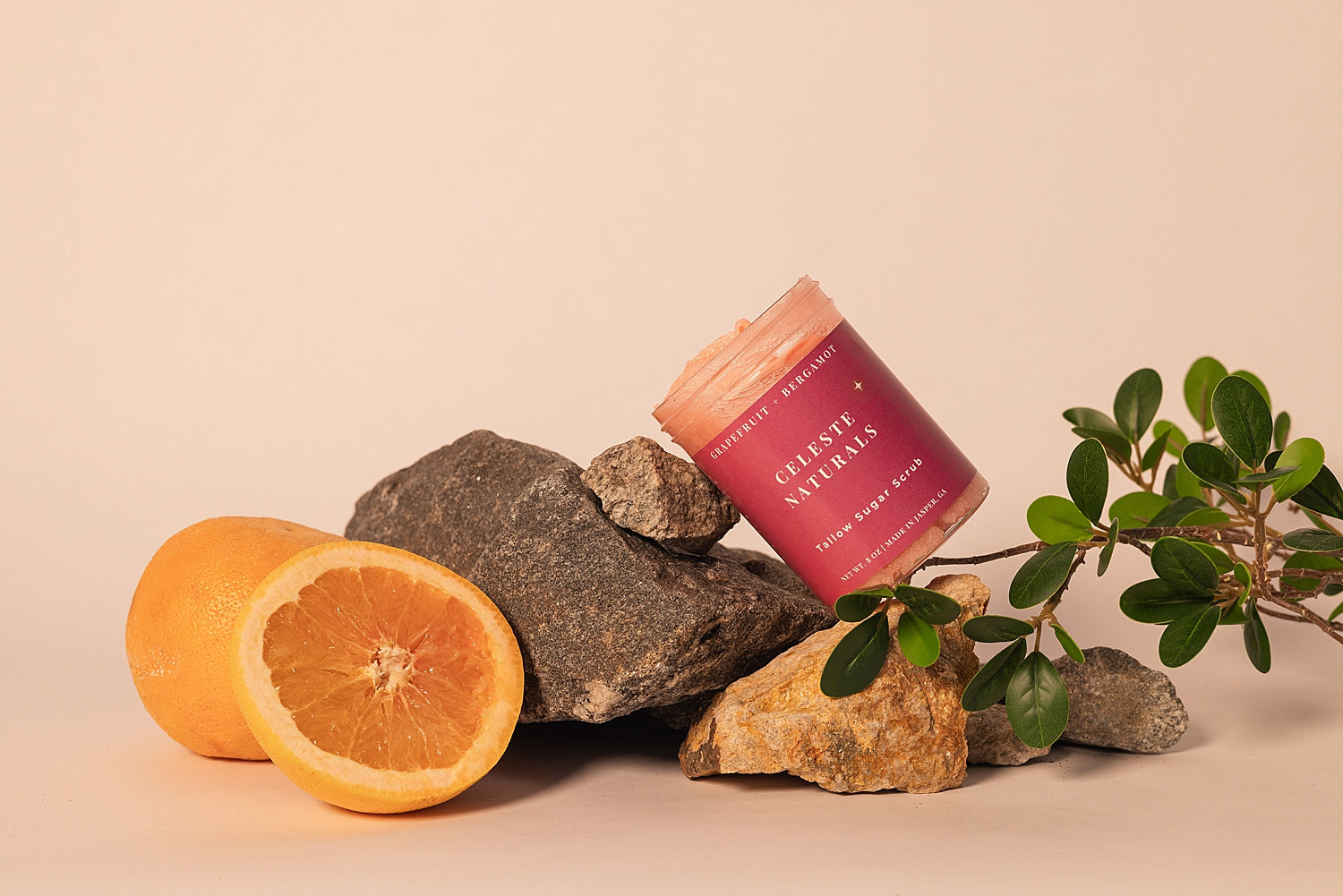 Jar of CELESTE NATURALS GRAPEFRUIT + BERGAMOT sugar scrub with oranges, rocks, and a plant on a beige background