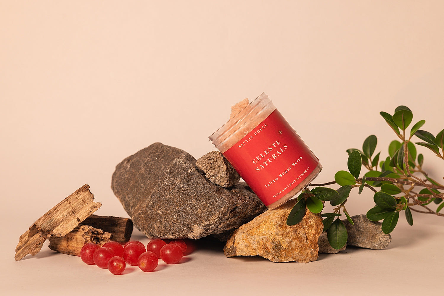 Jar of CELESTE NATURALS SANTAL ROUGE sugar scrub with wooden wick on rocks and berries against a beige background